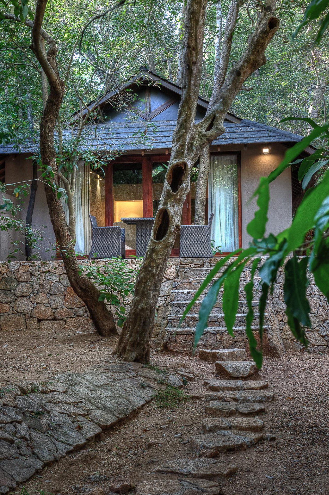 A cottage on a stone base among trees, with chairs on its terrace.