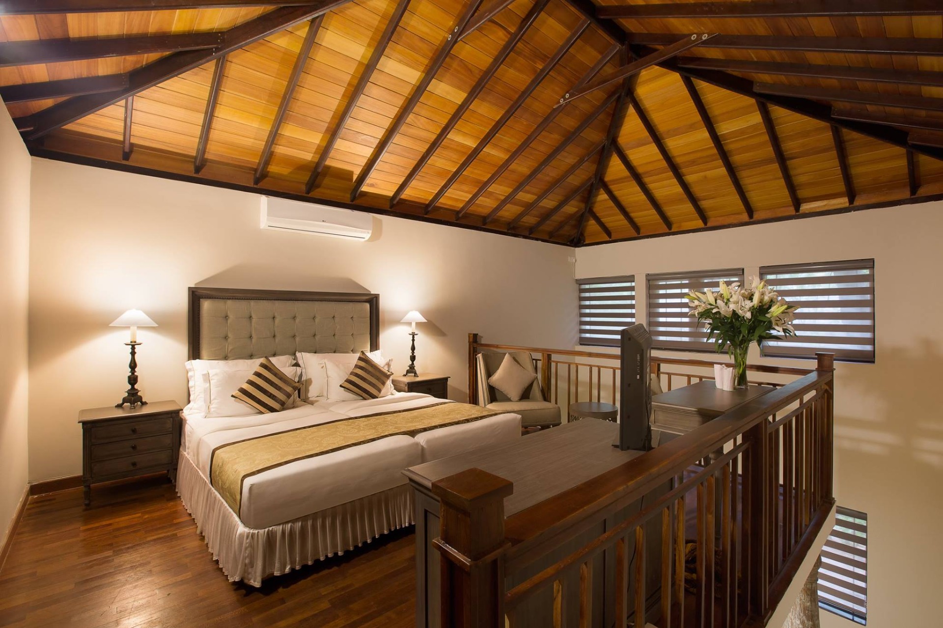 A bedroom beneath a timber roof, with a wooden balustrade and a bouquet on the side table.