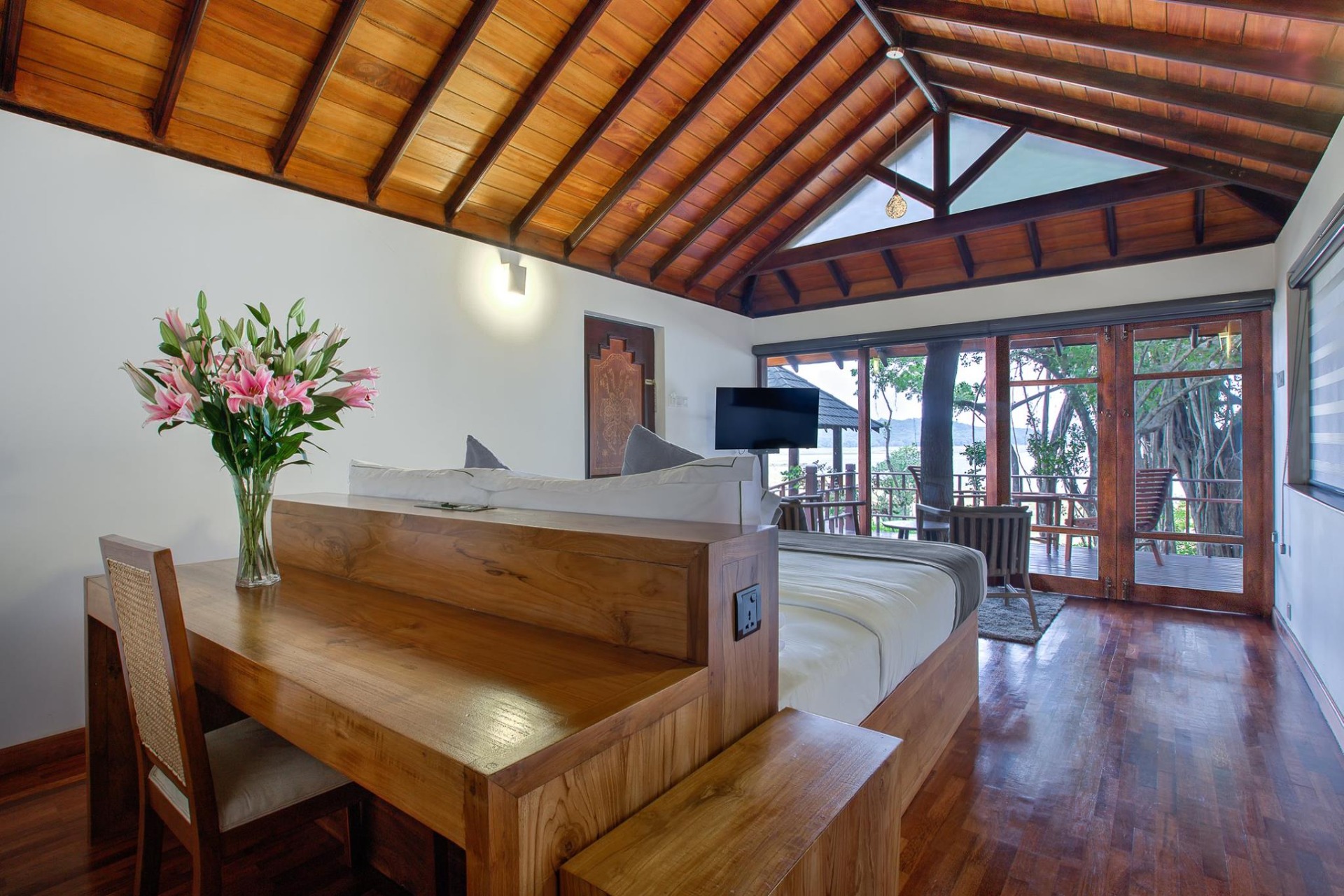 A room under a vaulted timber ceiling, with a bed, a writing desk and glass doors to the trees.